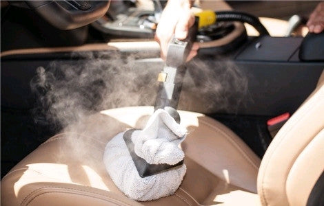  A hand-held steam cleaner with a white cloth attachment is being used to deep-clean a tan leather car seat. Steam is rising from the seat as it's being sanitized.
